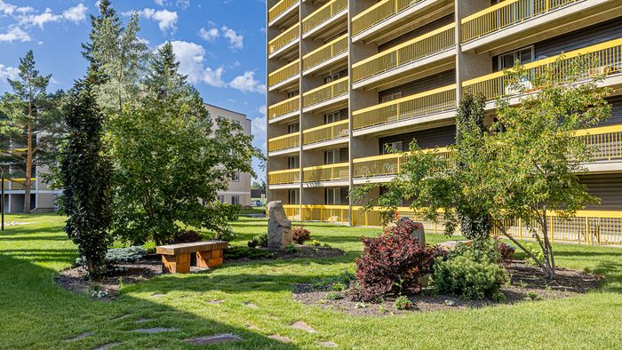 Charming apartment building exterior with vibrant yellow balconies overlooking a lush, green garden area with colorful plants and a rustic wooden bench.