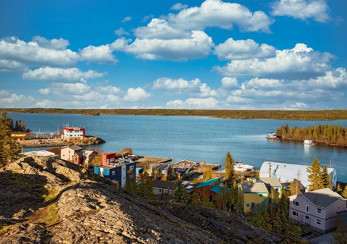 Scenic waterfront community with colorful homes and lush greenery overlooking a tranquil lake under a bright blue sky with fluffy clouds.