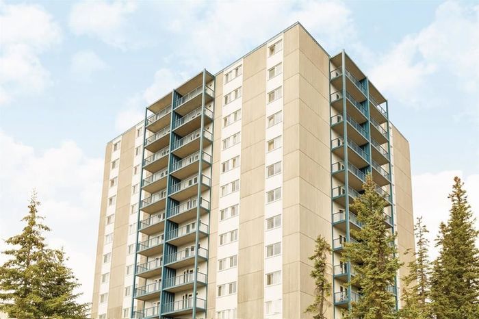 Modern high-rise apartment building with spacious balconies set against a clear blue sky and surrounded by lush green trees.