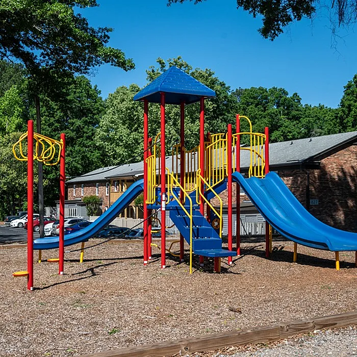 Colorful playground equipment set in a lush apartment complex, featuring slides and climbing structures surrounded by trees.