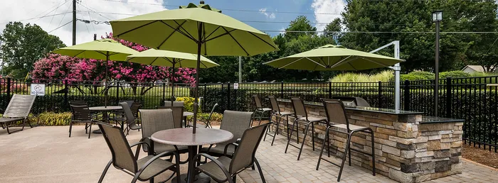 Chic outdoor patio with stylish stone bar, shaded by vibrant green umbrellas, surrounded by lush landscaping and blooming pink trees, perfect for relaxation and entertaining.