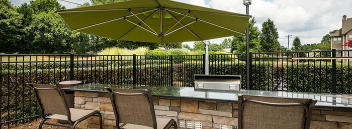 Charming outdoor patio with modern bar seating and sleek stone counter under a vibrant green umbrella, offering a relaxing view of lush greenery.