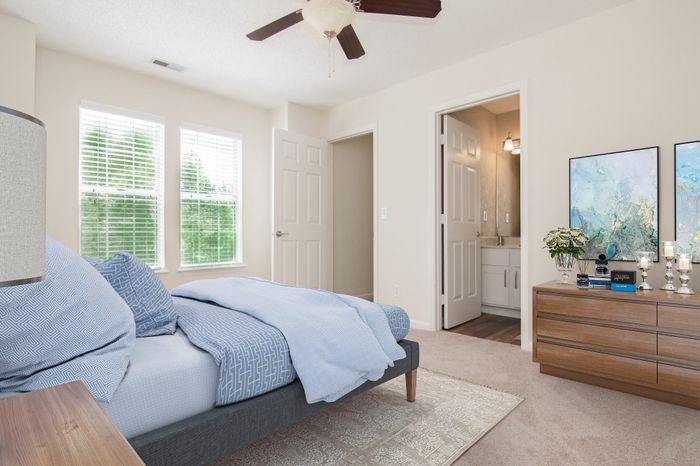 Bright and inviting bedroom with a cozy blue bed, elegant dresser, and soft carpet, enhanced by natural light from large windows.