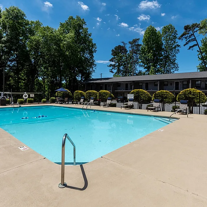 Inviting apartment complex outdoor pool with lush greenery and plenty of sunbathing chairs, perfect for relaxation under a clear blue sky.