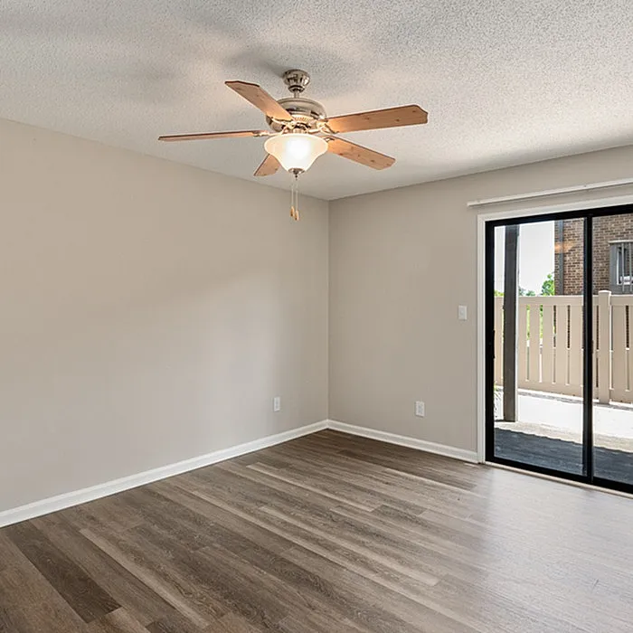 Bright, spacious room with sleek wooden flooring and modern ceiling fan, opening to a private patio through sliding glass doors.