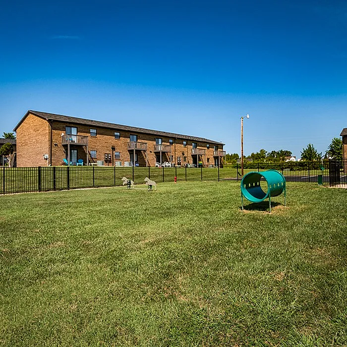 Charming apartment building with private balconies, set in a lush green outdoor area featuring a secure fenced yard and inviting play amenities.