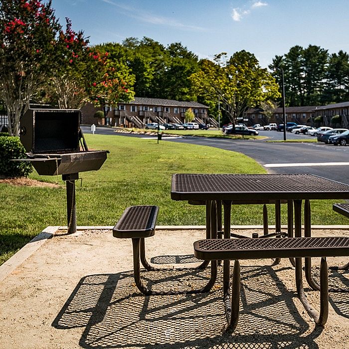 Charming outdoor area featuring a picnic table and grill, perfect for gatherings, set against a backdrop of lush greenery and apartment buildings.
