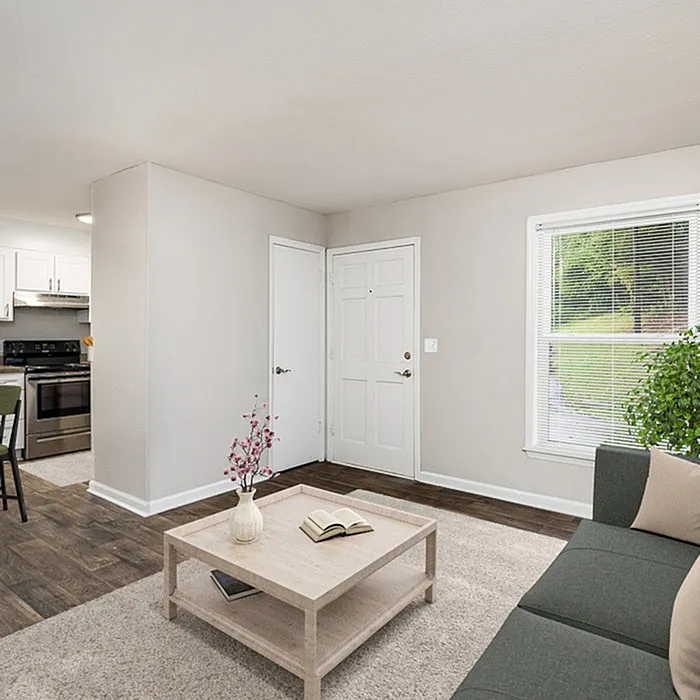 Bright and inviting living area featuring a cozy gray sofa, elegant coffee table with decor, and a view of lush greenery through the window, adjacent to a modern kitchen with stainless steel appliances.