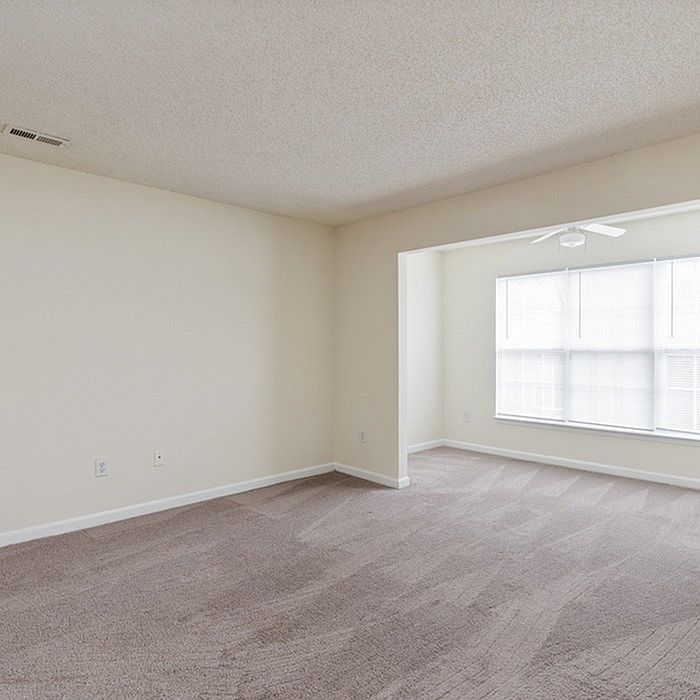 Bright and airy living space with plush carpeting, large windows, and modern ceiling fan, perfect for comfortable living.