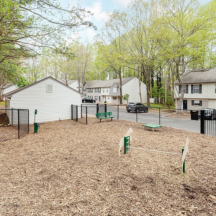 Charming apartment community features a cozy fenced yard with benches, surrounded by lush green trees and attractive residential buildings.