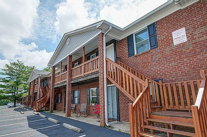 Charming brick apartment complex with wooden staircases, inviting balconies, and ample parking, surrounded by lush greenery under a vibrant sky.