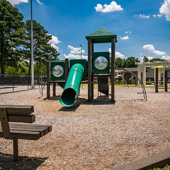 Charming play area with green slides and climbing structures, set against a sunny blue sky and surrounded by lush greenery.