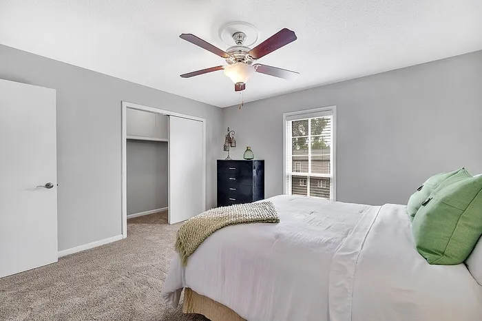 Bright and welcoming bedroom with soft neutral walls, featuring a cozy bed adorned with green pillows, a modern ceiling fan, and ample natural light streaming through a large window.
