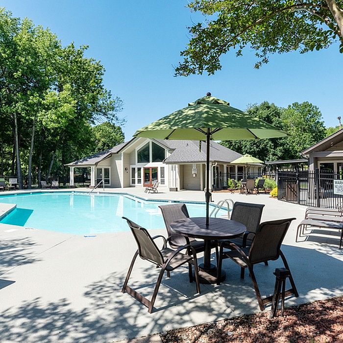 Inviting apartment complex with a sparkling pool, lush greenery, and cozy outdoor seating under a sun umbrella.