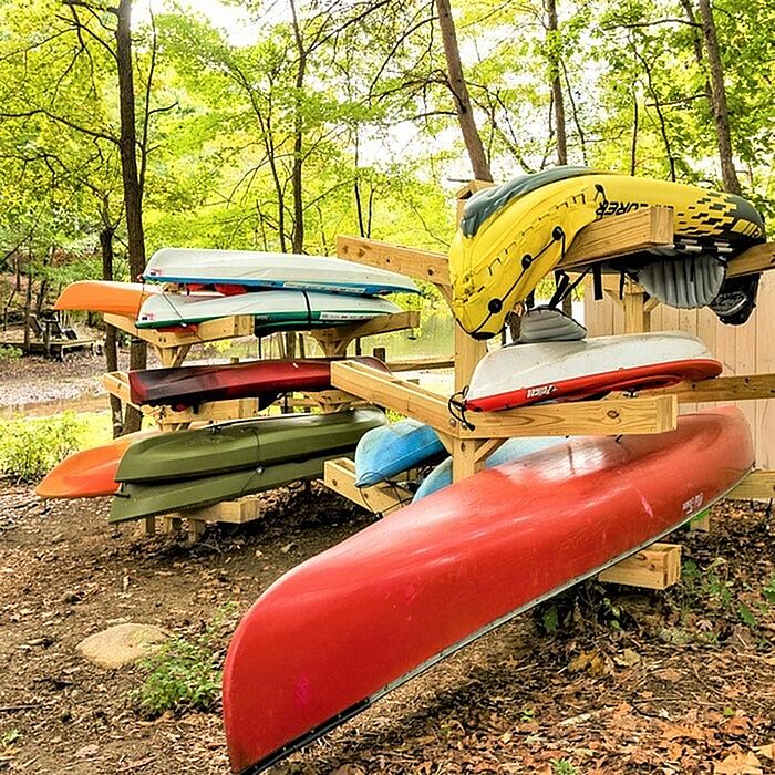 Vibrant collection of kayaks and canoes neatly stacked on wooden racks, set against a lush, green forest backdrop.