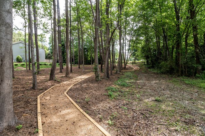 Charming wooded trail leading to a secluded residential area, surrounded by lush greenery and tall trees, perfect for a peaceful nature walk.