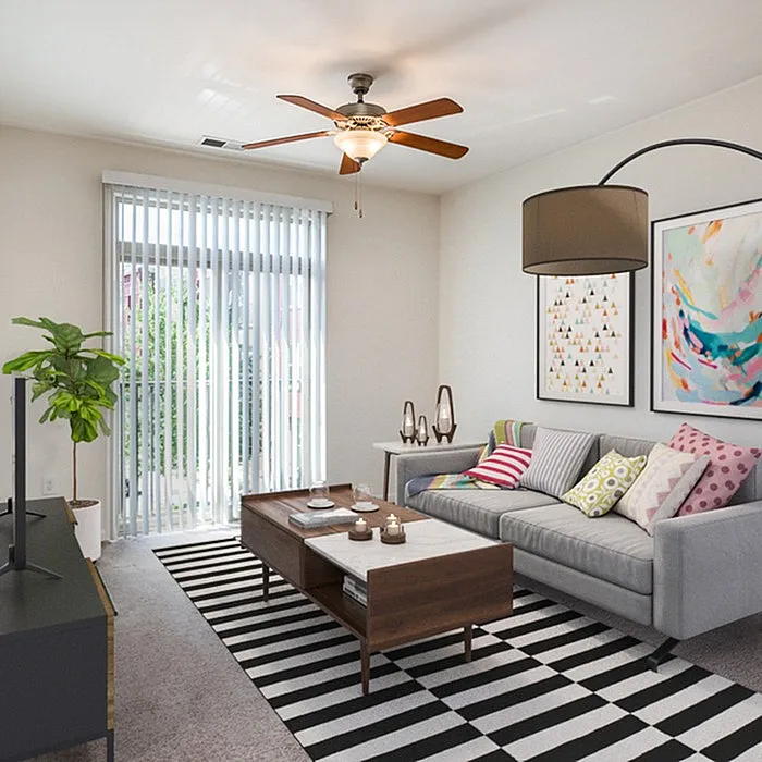 Bright and inviting living room featuring a cozy gray sofa with colorful cushions, modern artwork, and a stylish striped rug. Abundant natural light streams through large sliding glass doors with vertical blinds, complemented by a chic ceiling fan.