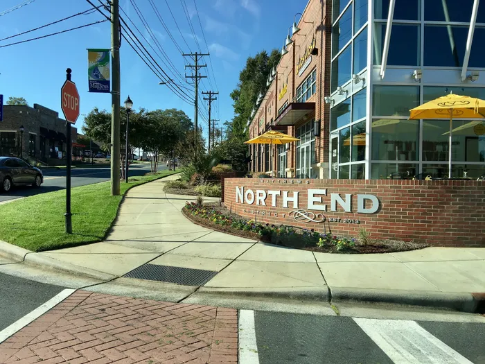 Charming North End district with modern brick design, sidewalk cafes with umbrellas, and lush greenery, creating an inviting community atmosphere.