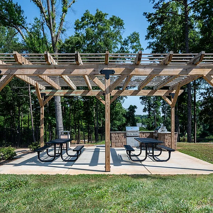 Inviting outdoor area with wooden pergola, picnic tables, and modern grill set against a lush, green backdrop.