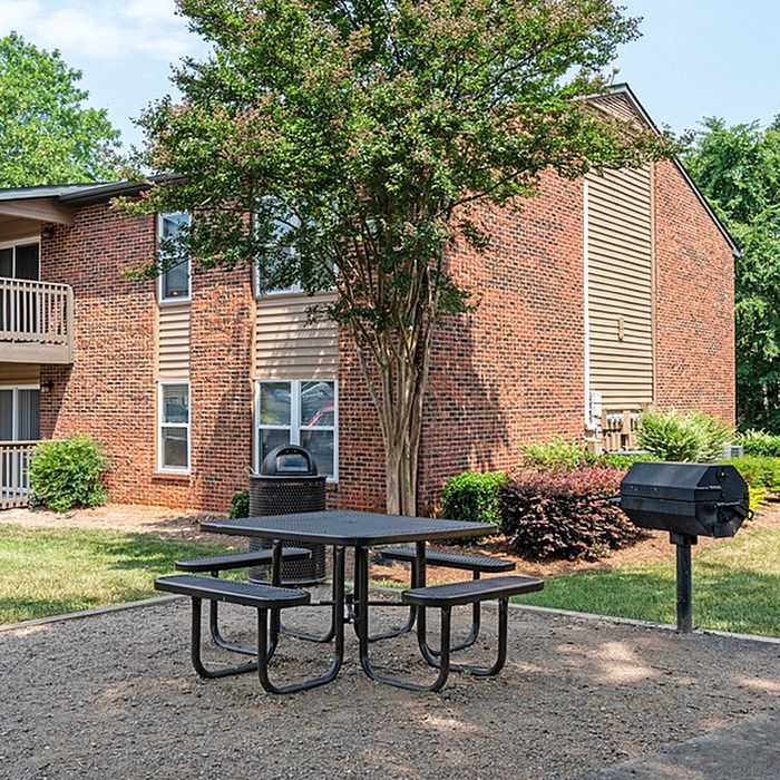 Charming apartment exterior with a cozy brick facade, complemented by a lush tree and a convenient outdoor picnic area with a barbecue grill.