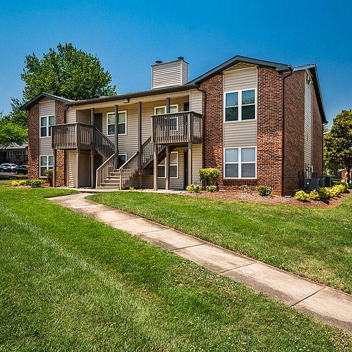 Charming two-story apartment building with brick facade and balcony, surrounded by lush greenery and a well-maintained lawn.