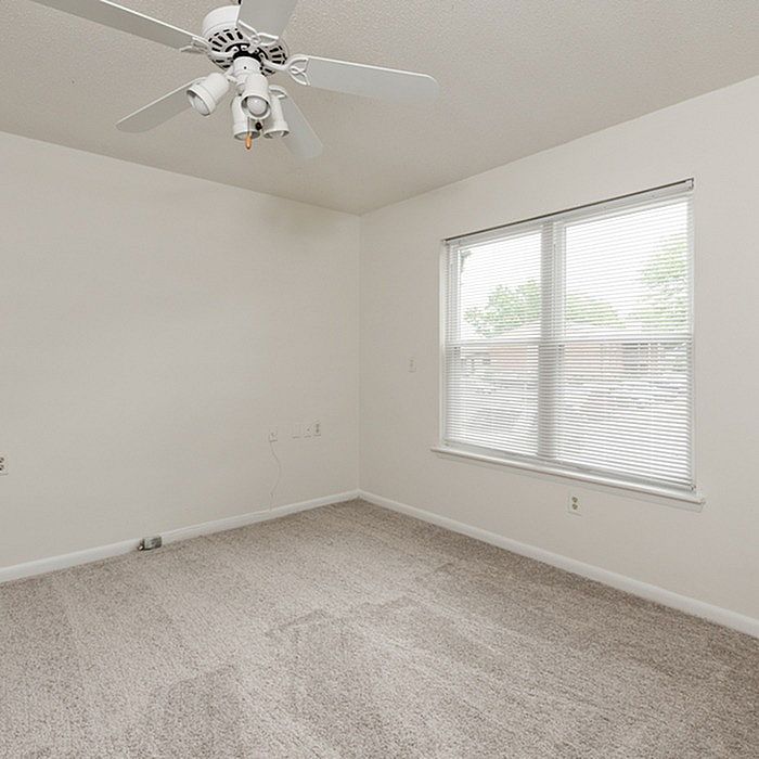 Bright, airy bedroom with large window and ceiling fan, featuring plush carpeting and a neutral color palette for a cozy ambiance.