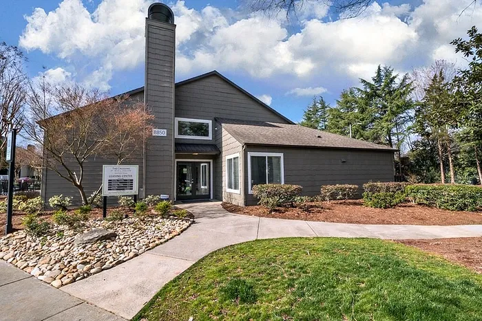 Charming leasing center with modern design surrounded by lush landscaping and a peaceful walkway under a bright blue sky.