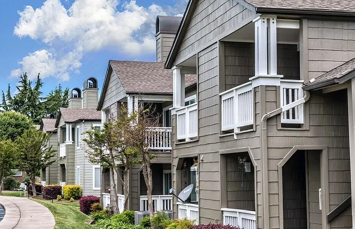 Charming residential apartment complex with quaint wooden balconies, lush greenery, and a serene pathway under a bright blue sky.