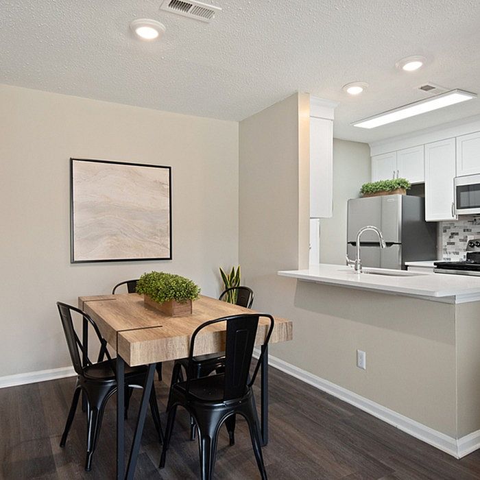 Modern apartment dining and kitchen area featuring a sleek wooden table with black chairs, contemporary white cabinets, and stainless-steel appliances.