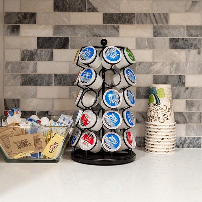 Modern kitchen countertop featuring a stylish coffee pod carousel, decorative paper coffee cups, and a bowl of assorted sweeteners against a chic tile backsplash.