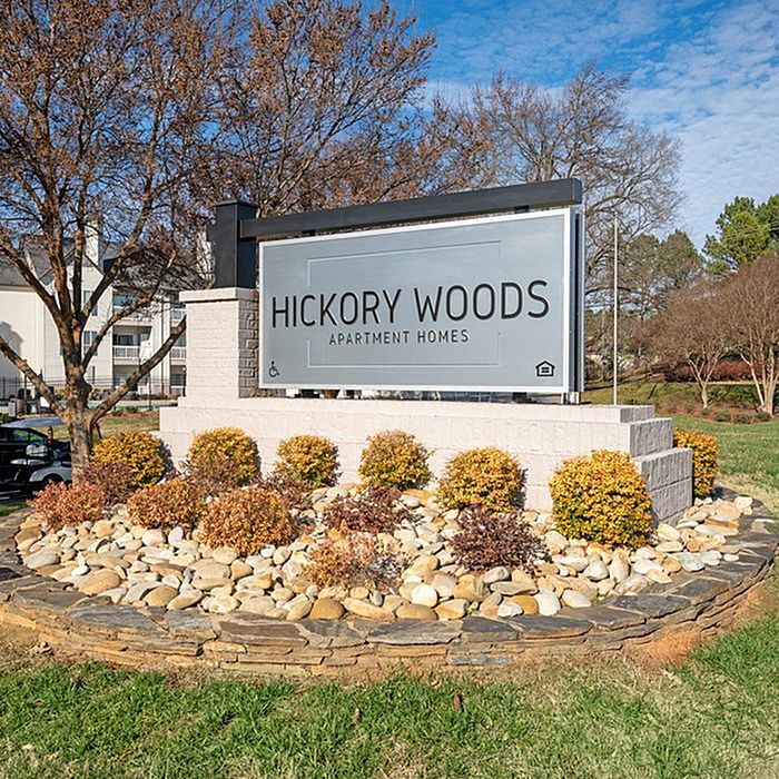 Inviting entrance sign for Hickory Woods Apartment Homes, featuring landscaped stone and shrubbery, set against a backdrop of trees and a blue sky.
