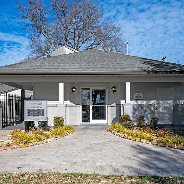 Charming leasing center with a welcoming facade, surrounded by landscaped greenery and a paved entrance, under a bright blue sky.
