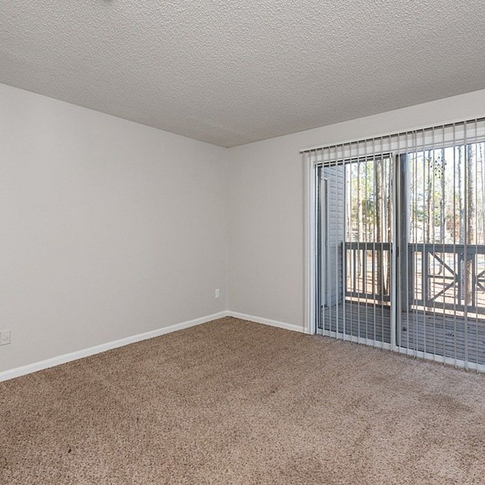 Bright apartment room with plush carpet and large sliding glass doors opening to a serene private balcony.