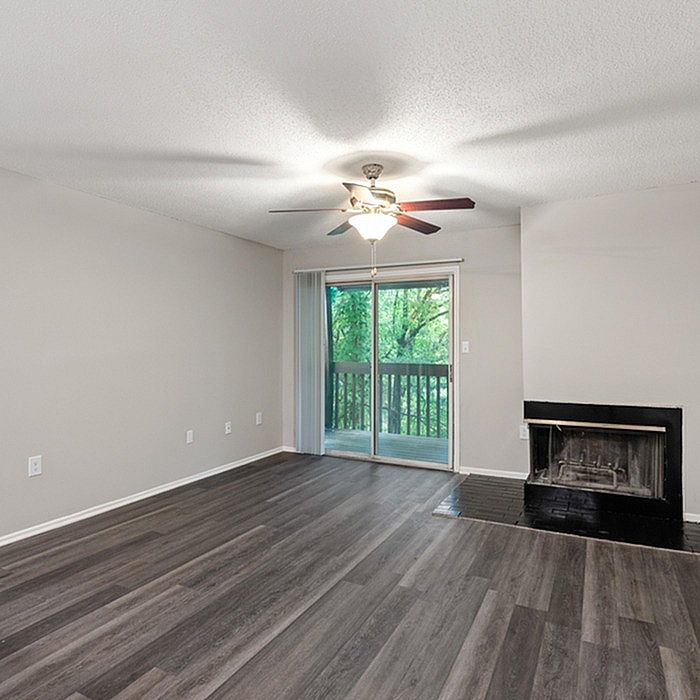 Bright and airy living room featuring sleek wood flooring, a modern ceiling fan, and a cozy fireplace, with a view of lush greenery through sliding glass doors.