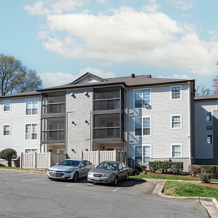 Modern apartment complex with three-story structure, multiple balconies, ample parking, and scenic greenery under a bright blue sky.
