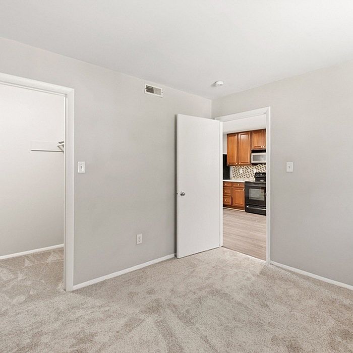 Bright and inviting apartment room with plush carpeting, leading to a modern kitchen featuring wooden cabinets and sleek appliances.
