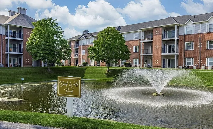 Charming apartment complex with red brick exteriors overlooking a serene pond with a beautiful fountain and lush greenery.