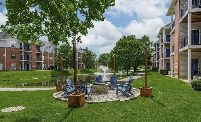 Charming apartment complex with a vibrant green courtyard featuring Adirondack chairs around a fire pit, flanked by lovely trees and a serene pond with a fountain.