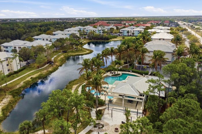Aerial view of a luxurious apartment complex with a stunning pool, surrounded by lush greenery and a serene lake, offering a tropical oasis lifestyle.