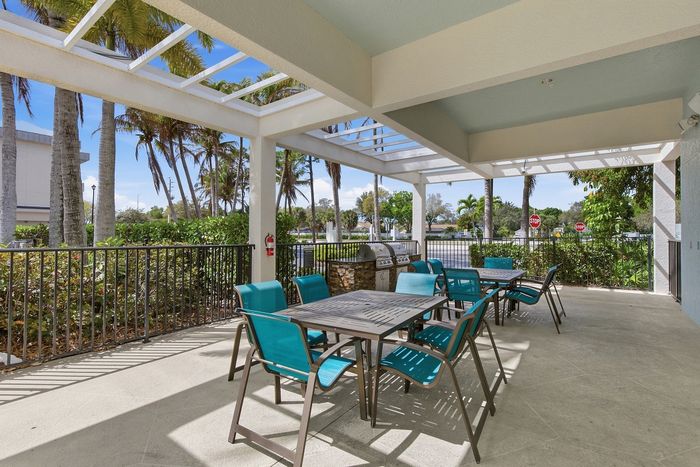 Charming outdoor patio with modern blue seating, shaded pergola, and built-in BBQ grill offering a relaxing, tropical vibe surrounded by palm trees.