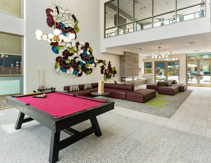 Vibrant and modern lounge with a stylish billiard table, cozy maroon sofas, eye-catching art installation, and floor-to-ceiling windows offering abundant natural light.