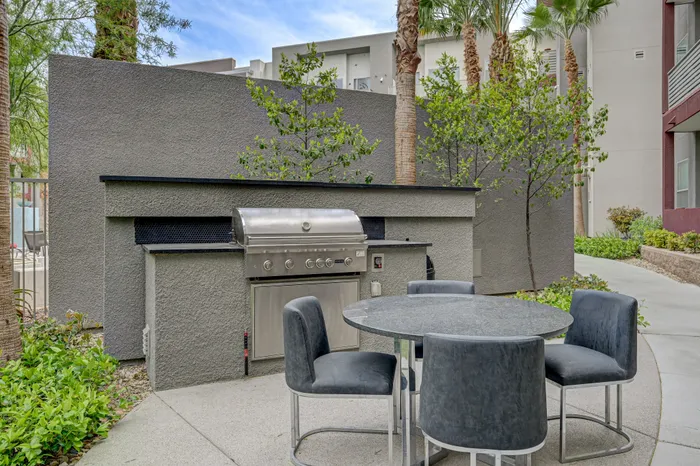 Chic outdoor patio area featuring a modern stainless steel grill and stylish round dining table with seats, surrounded by lush greenery and palm trees.