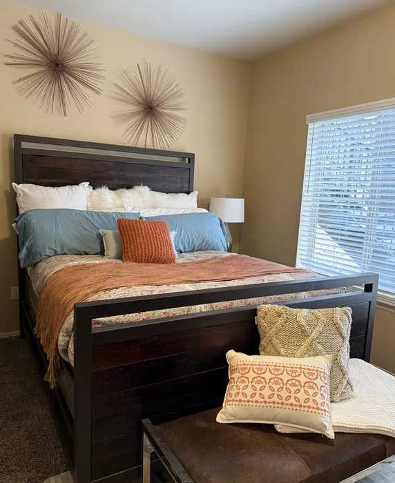 Cozy bedroom featuring a plush bed with vibrant pillows and modern wall decor, inviting natural light through large window blinds.