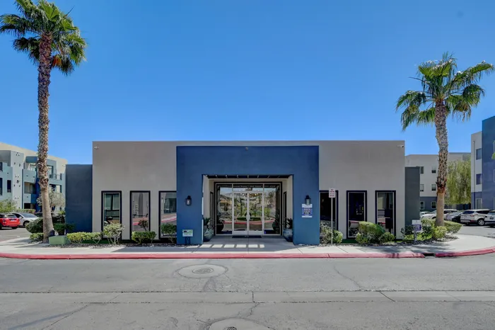 Modern apartment complex entrance with sleek architecture, lush landscaping, and palm trees under a clear blue sky.