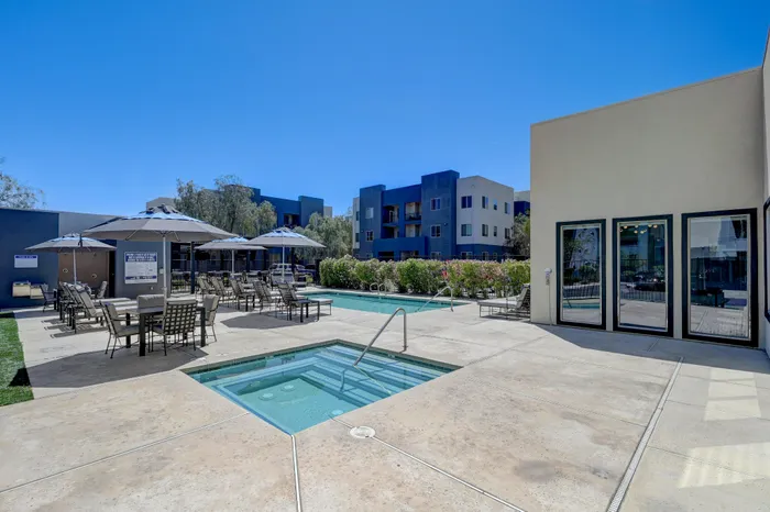 Modern apartment complex with luxurious outdoor pool and spa area, surrounded by comfortable lounge chairs and umbrellas under a clear blue sky.