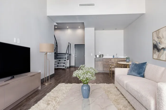 Bright and modern living room with a cozy couch, sleek flat-screen TV, and stylish decor. Features a contemporary kitchen area and elegant staircase leading to a loft space.