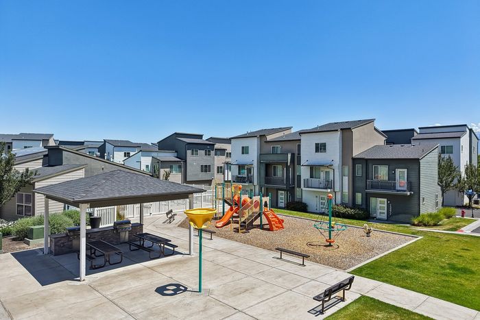 Vibrant apartment complex featuring modern units with private balconies, a playful kids' playground, and a communal BBQ area under a sunny blue sky.