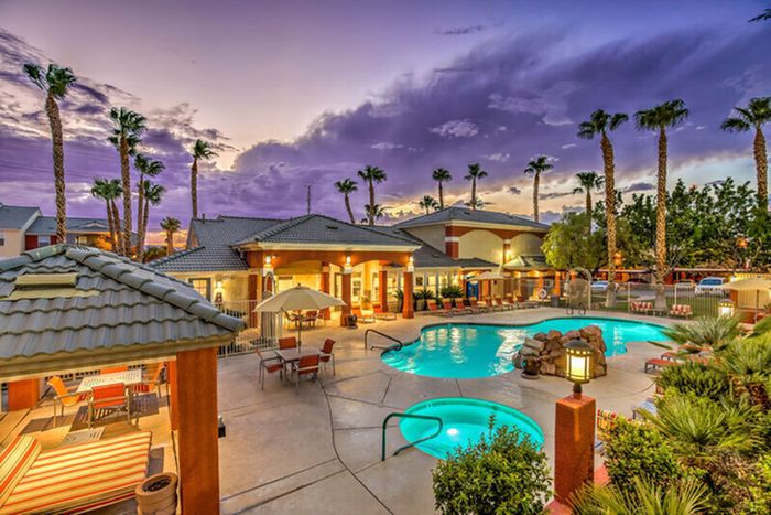 Luxurious apartment complex with a sparkling pool and inviting outdoor lounge area under a stunning sunset sky, bordered by elegant palm trees.
