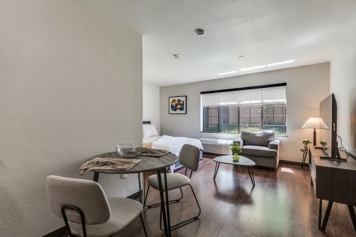 Bright and inviting studio apartment with modern furnishings, featuring an open layout, cozy sitting area, and large window letting in natural light.