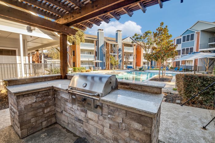 Outdoor grilling station with stainless steel barbecue and view of resort-style pool surrounded by modern apartment buildings at Preferred Place Houston.